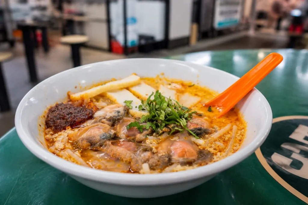 Bowl of spicy noodle soup with cockles and an orange spoon
