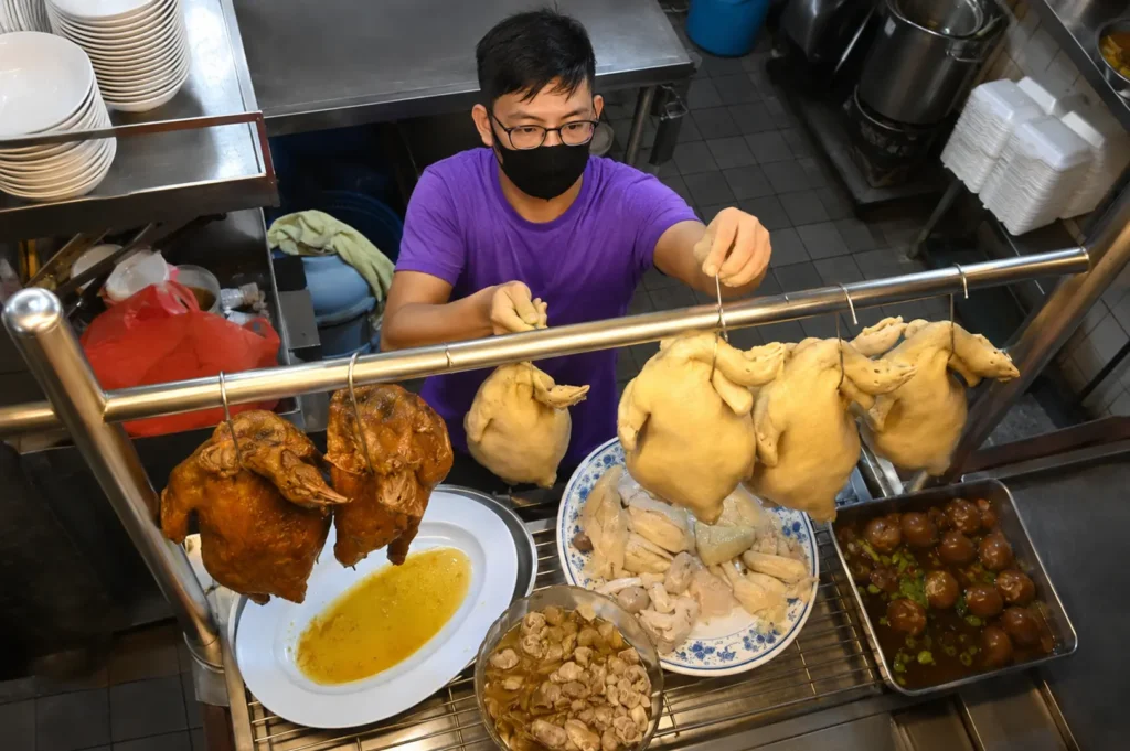 Hawker worker preparing whole roasted and poached chickens on a rack with braised eggs and chopped chicken