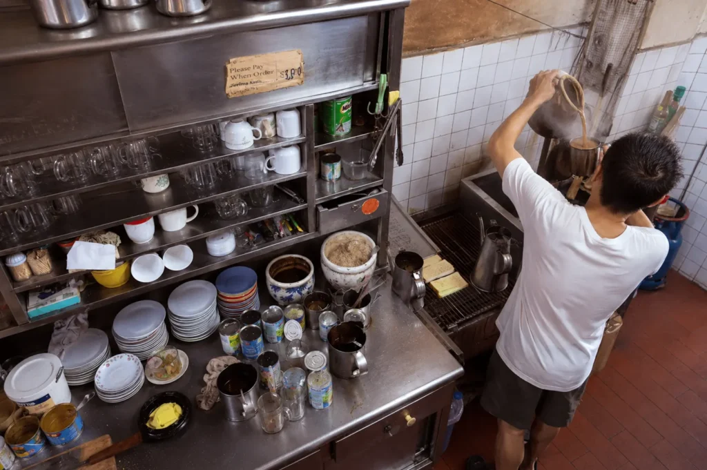 Kopitiam worker preparing traditional coffee at stainless steel beverage station with stacked cups