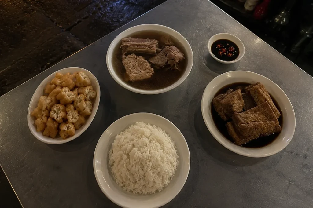 Bowls of bak kut teh, braised tofu, fried dough fritters, and rice on a metal table at a late-night supper spot