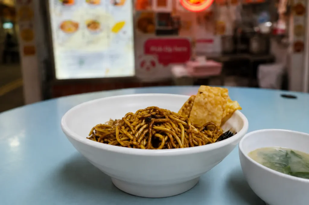 Bowl of dry hawker noodles with fried wonton and side soup on a blue table