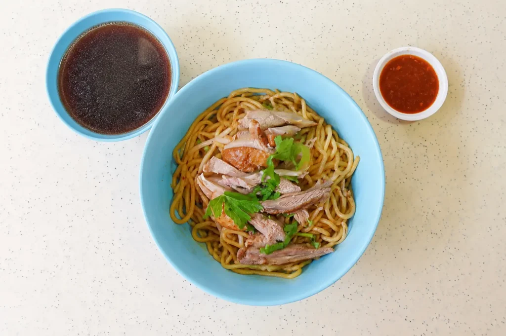 Top-down view of a blue bowl filled with dry noodles topped with sliced meat and cilantro, served with a side of dark broth and chili sauce on a speckled hawker table