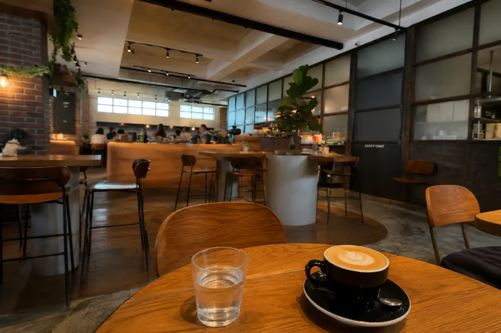 Black cup of latte and water glass on a wooden table, with the spacious industrial interior of Five Oars Coffee Roasters in the background