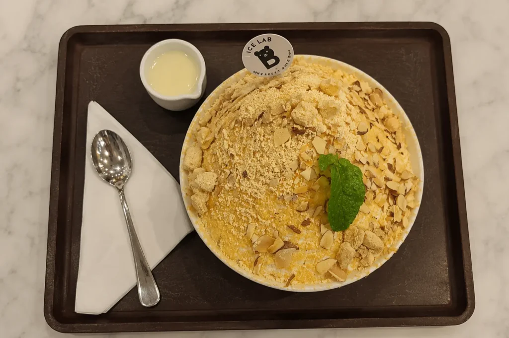 Top-down view of a large bowl of bingsu topped with crumbled cookies, nuts, and a mint leaf, served on a dark tray with condensed milk