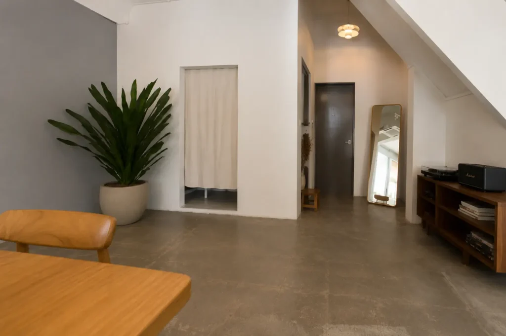 Minimalist apartment-style cafe interior featuring a wooden table, large potted plant, and a cozy corner