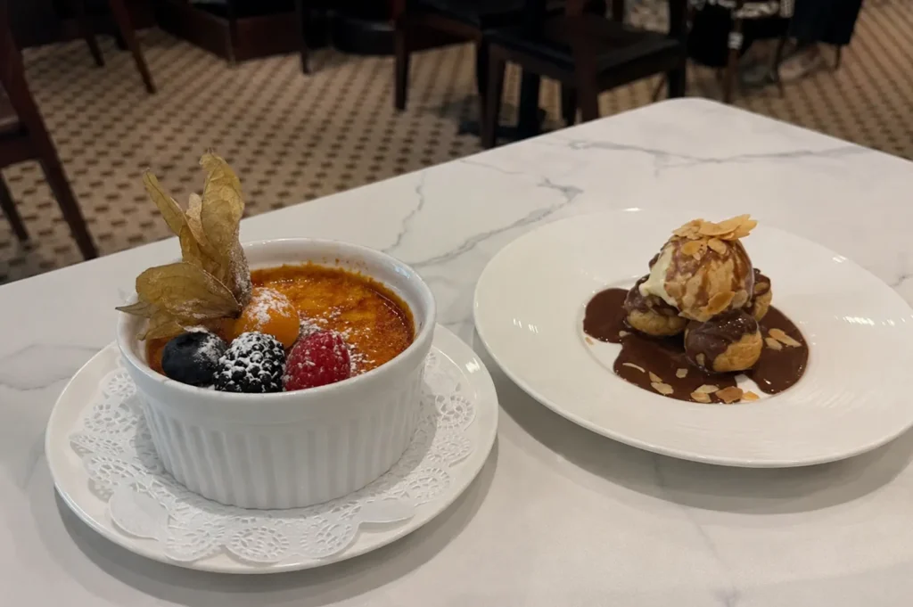 Two French desserts on a marble table featuring a berry-topped crème brûlée and profiteroles with chocolate sauce