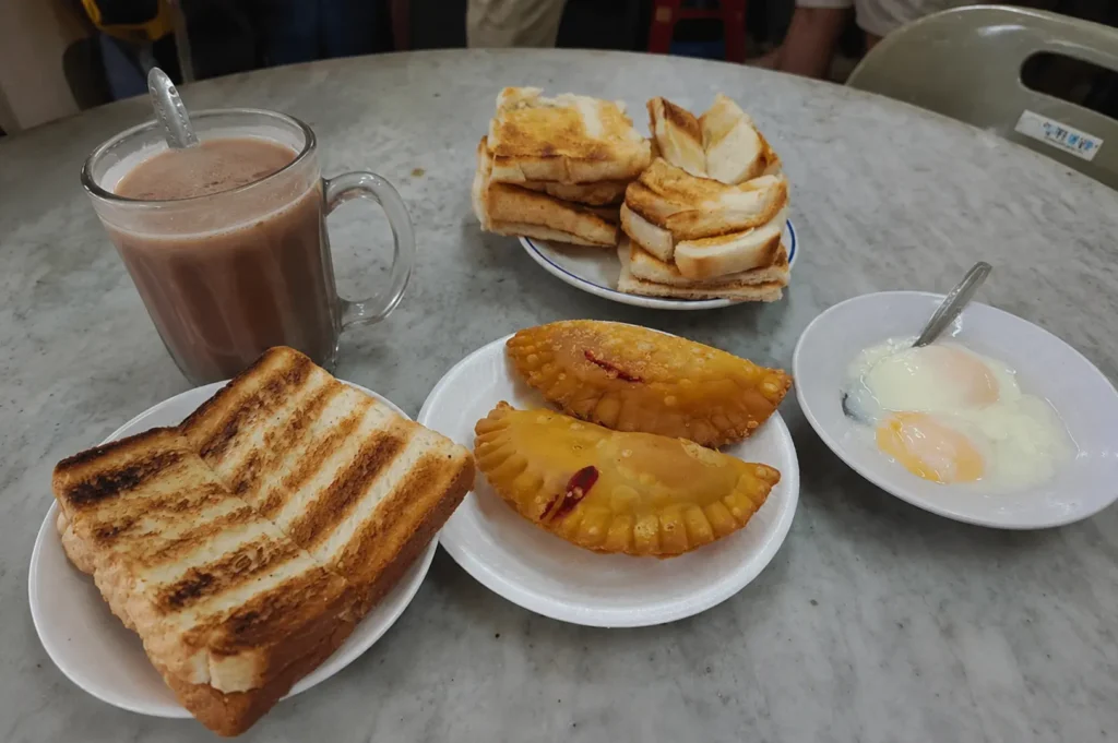 Full kopitiam breakfast spread with kaya toast, curry puffs, soft-boiled eggs and milky tea