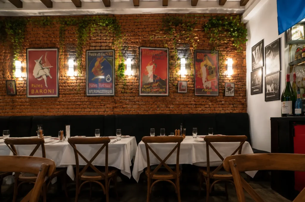 Interior view of a French bistro featuring an exposed brick wall adorned with vintage posters and ivy, warm wall lighting, and dining tables set with white tablecloths and wooden chairs