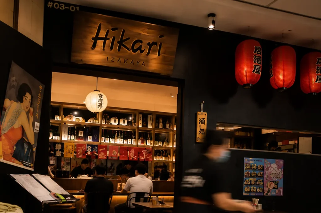 Hikari Izakaya entrance with wooden sign, red lanterns, and shelves of sake bottles