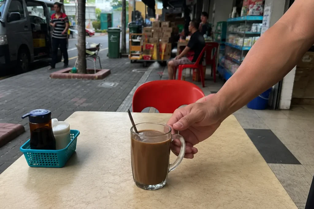 Hand holding glass mug of traditional kopi at outdoor kopitiam table with condiment basket and red plastic chairs