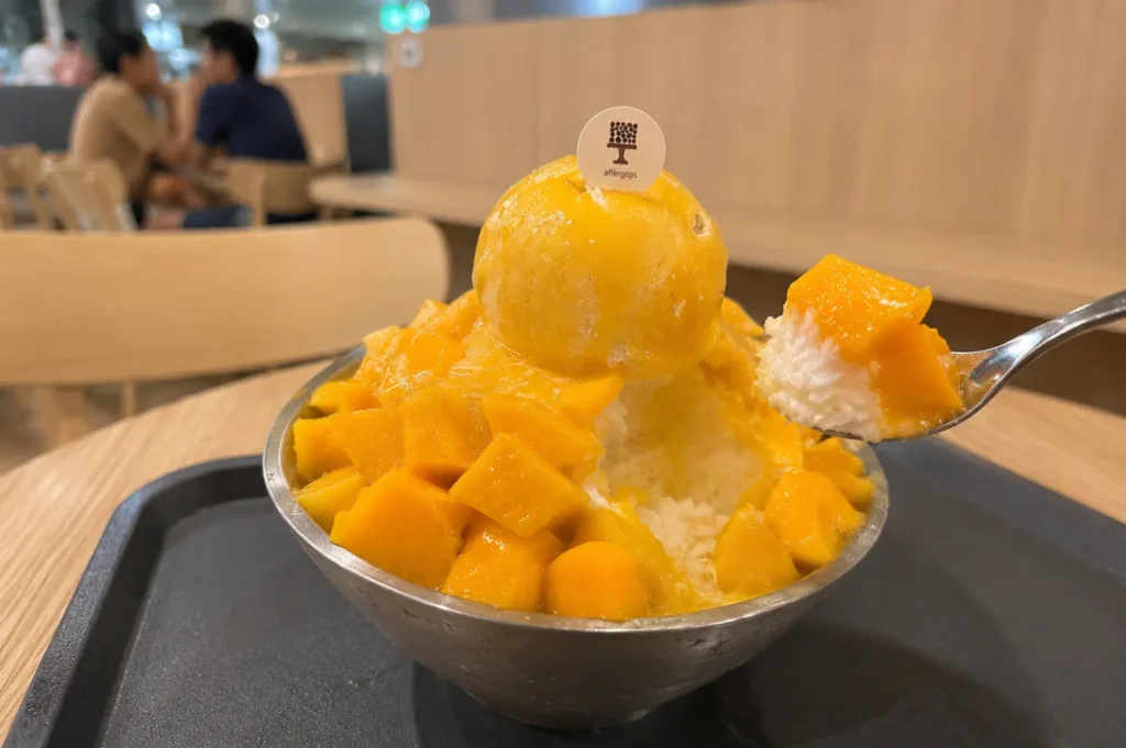 Bowl of mango bingsu topped with fresh mango cubes and a scoop of sorbet, with a spoon lifting a bite