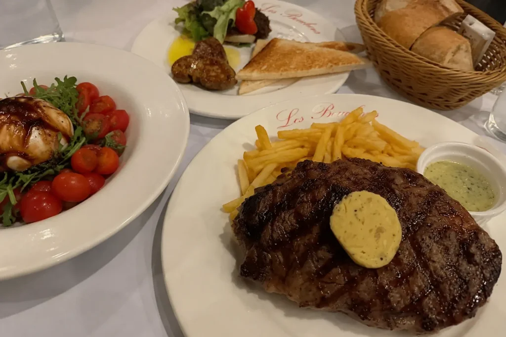 Close-up of a grilled steak topped with herb butter served with french fries on a Les Bouchons branded plate, accompanied by a fresh tomato salad, foie gras appetizer, and a basket of bread