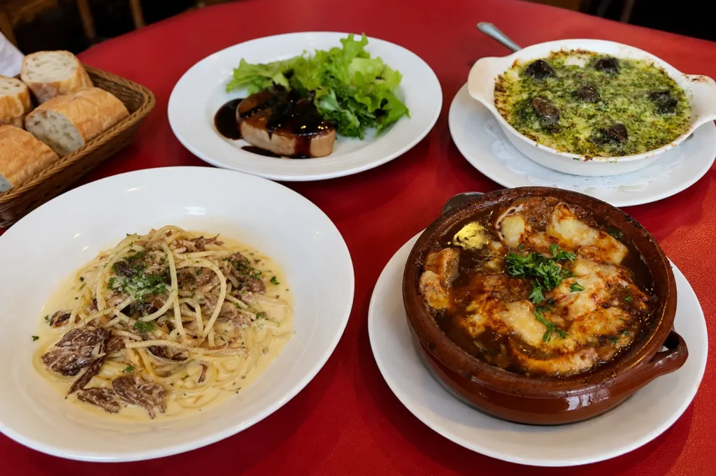 Spread of French dishes including onion soup, pasta, and foie gras on a red tablecloth
