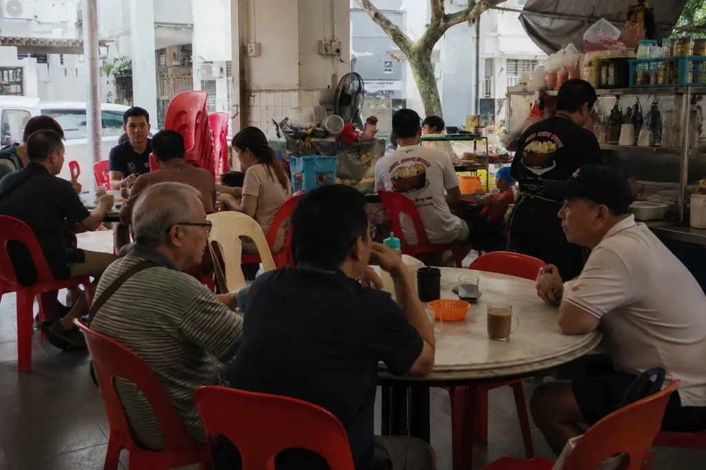 Busy kopitiam dining area with customers seated at tables with red plastic chairs in open-air setting