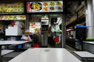 Chomp Chomp hawker stall interior with illuminated menu boards, stainless steel kitchen workspace, and empty tables before the dinner rush.