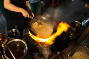 Chef stir-frying noodles over high flame at a Singapore hawker stall.