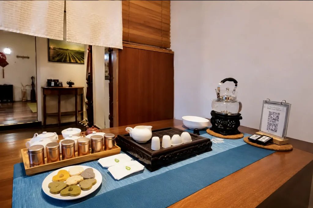 Traditional Chinese tea ceremony setup with white porcelain teapots, tasting cups, metal canisters, matcha cookies, and a glass kettle on a blue table runner
