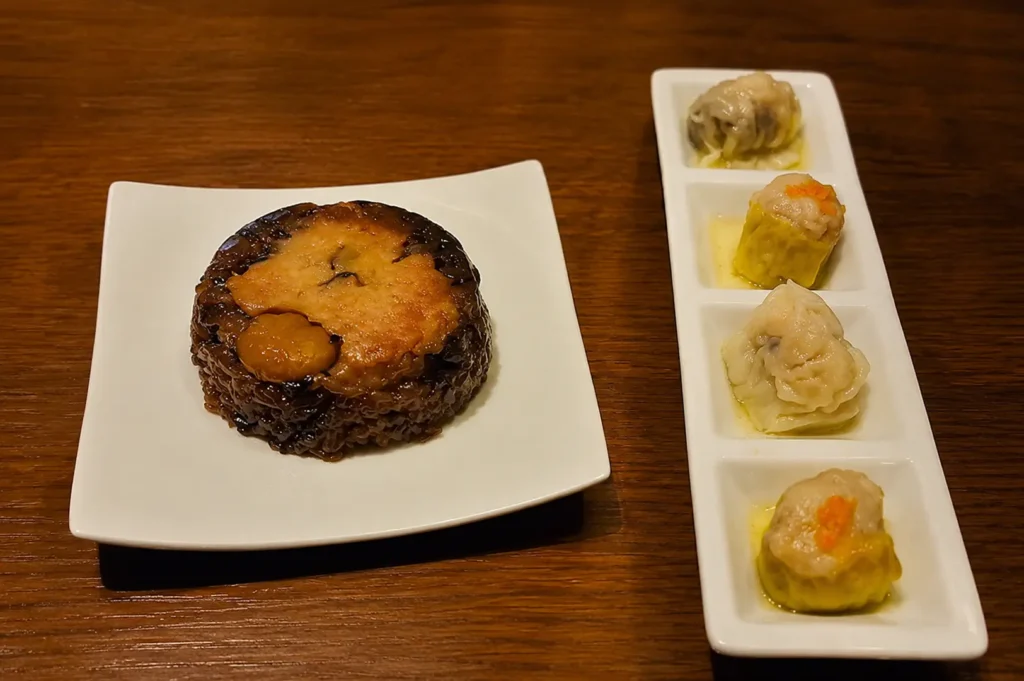 A molded dark sticky rice dish with toppings on a square plate, served alongside a four-compartment sampler of dim sum dumplings