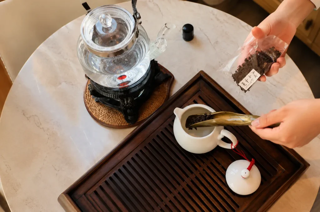 Server using a bamboo scoop to place loose tea leaves into a white teapot on a wooden tray, next to a glass kettle